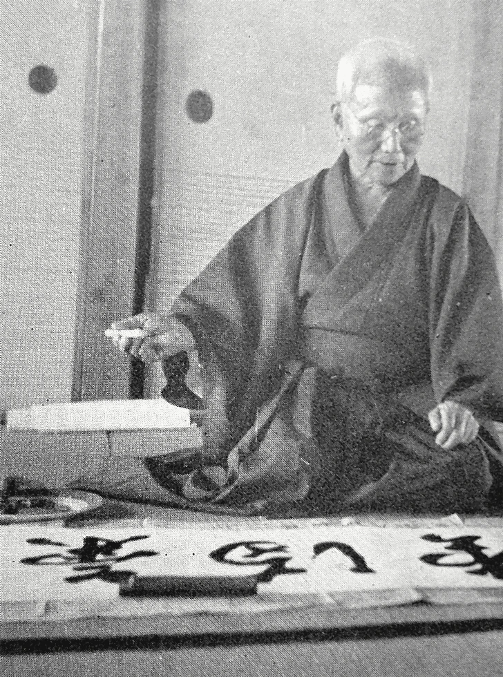 O-Sensei Gichin Funakoshi seated on tatami holding brush next to his calligraphy Ichiban