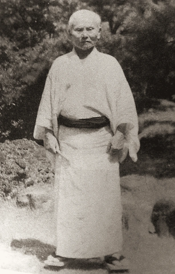 O-Sensei Gichin Funakoshi standing outdoors in traditional kimono and geta